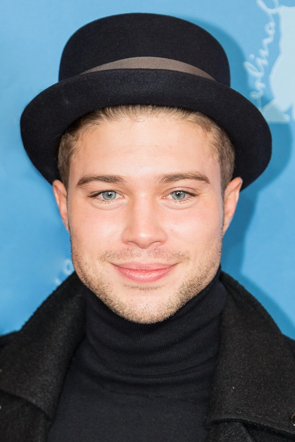 Portrait of a young man wearing a black hat and dark coat against a blue background