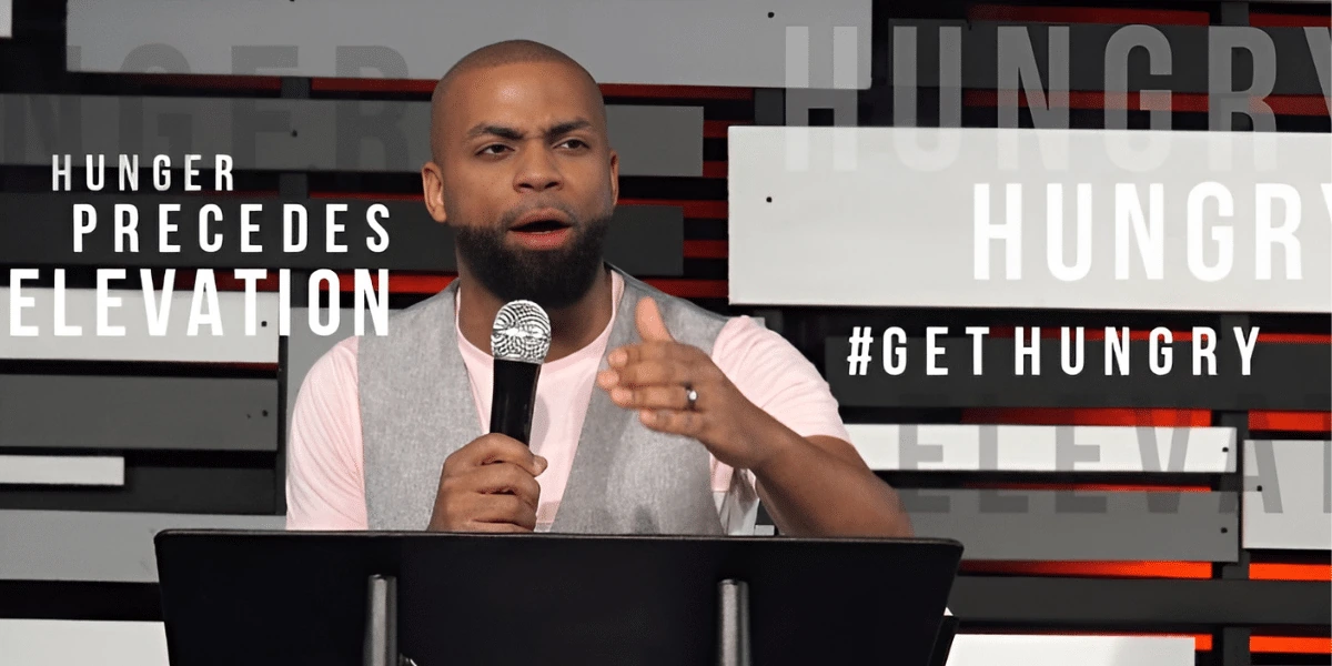 Marcus “Elevation” Taylor speaking on stage with a microphone in front of a “Hunger Precedes Elevation” backdrop.